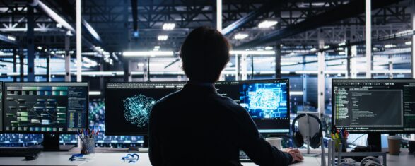 Silhouetted person seated at a multi-monitor desk in a dark server facility, working with AI neural network visualizations and a glowing brain-shaped diagram on the center screens, flanked by displays showing code, data dashboards, and network graphs.