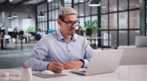 Man with glasses writing notes at a desk while reviewing content on a laptop in a modern office
