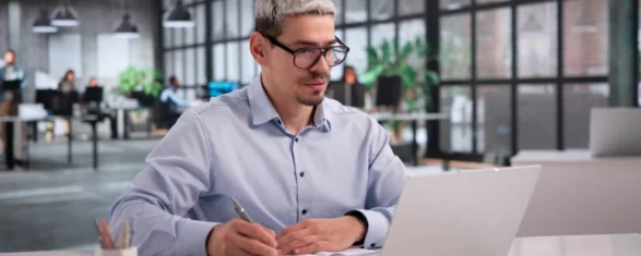 Ein Mann mit Brille, der an einem Schreibtisch Notizen macht, während er in einem modernen Büro Inhalte auf einem Laptop durchsieht