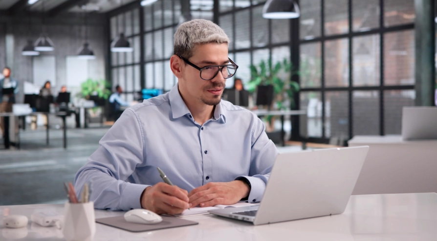 Ein Mann mit Brille, der an einem Schreibtisch Notizen macht, während er in einem modernen Büro Inhalte auf einem Laptop durchsieht