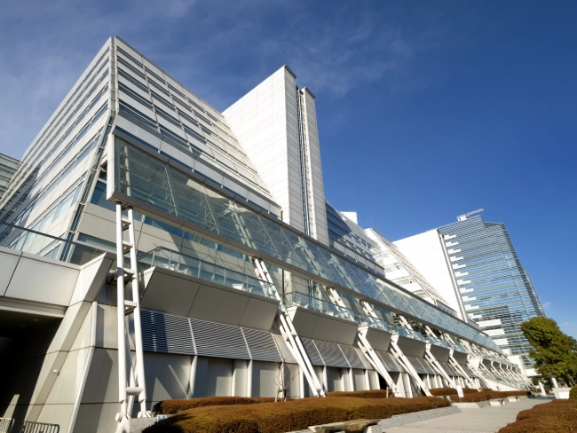 Exterior view of TFT Hall (Tokyo Fashion Town Building) in Tokyo, showing its angular white and glass facade under a clear blue sky, with city skyscrapers in the background