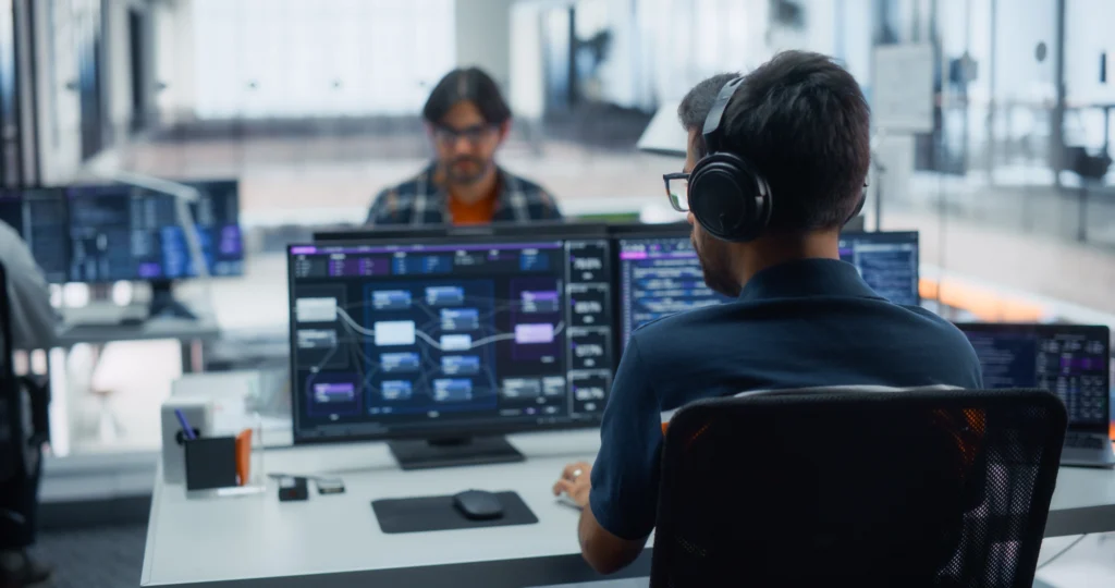 Engineer wearing headphones working at a workstation with workflow dashboards on two monitors in an open-plan office